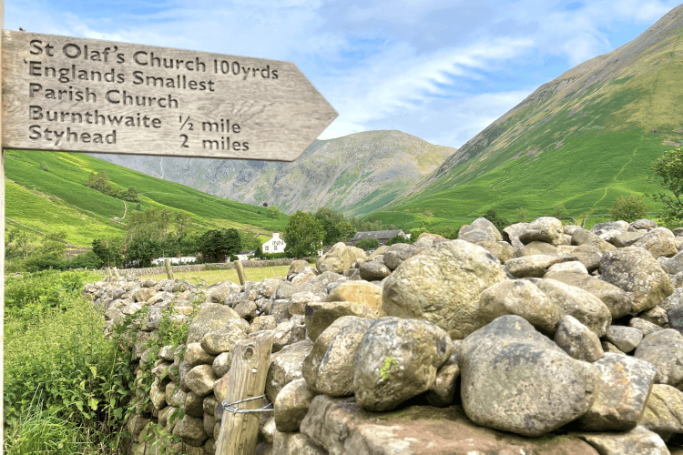 A wooden fingerpost in the foreground points out Olaf's Church, Burnthwaite and Styhead. Beyond the sign and a loose-stacked stone wall, a pub nestles at the crux of several green hills, with the bulk of a Lakeland fell rising in the far distance.